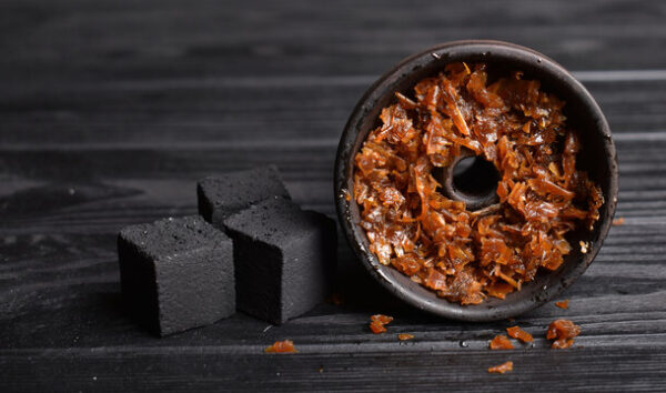 A shisha bowl filled with moist, shredded tobacco mixture placed beside three black charcoal cubes on a dark wooden surface.