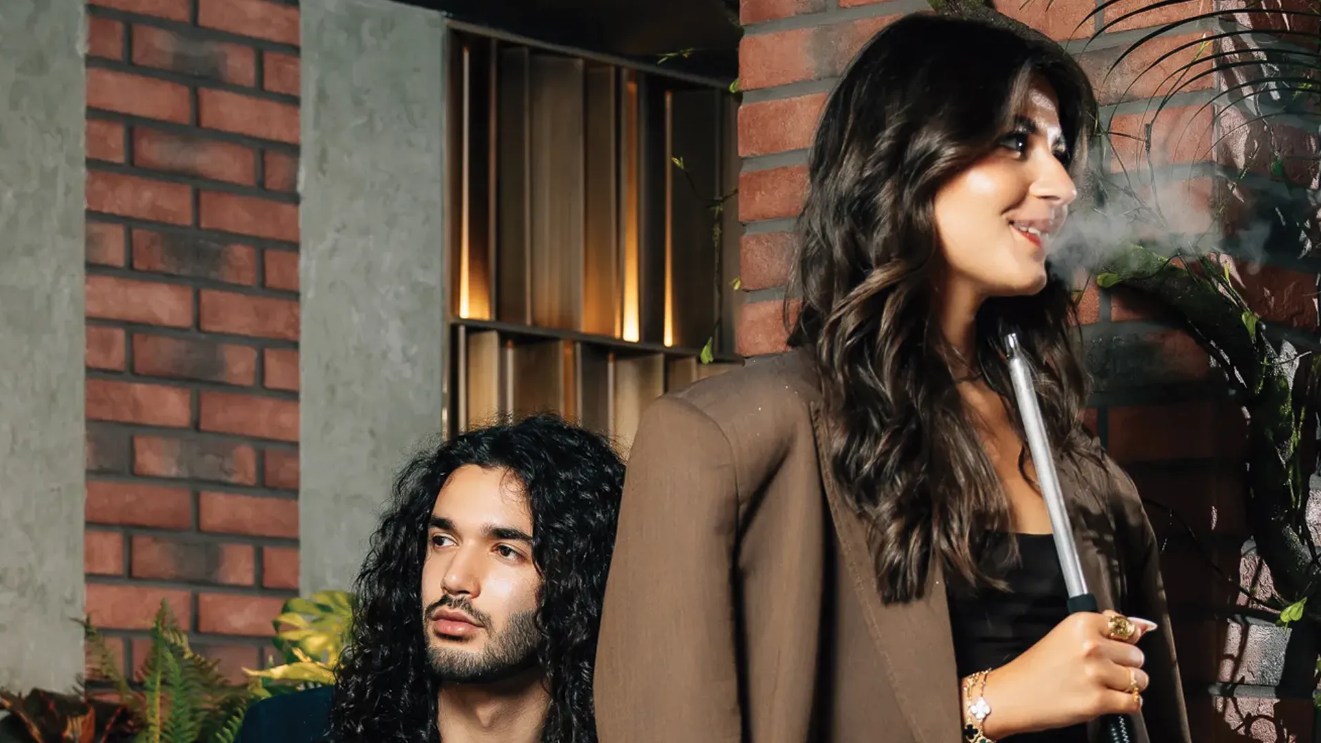 Two people relaxing outside a lounge, one holding a shisha pipe with a brick wall background.