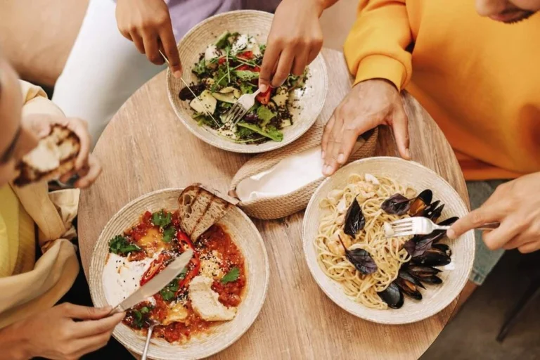 A top view of three people dining at a round wooden table, enjoying bowls of fresh salad, seafood pasta, and a tomato-based dish with bread, creating a warm and casual meal setting.