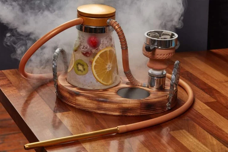 A modern fruit-infused shisha setup on a wooden table, featuring a glass base filled with ice, lemon slices, kiwi, and berries, with smoke rising around the hookah and its tan-colored hose.