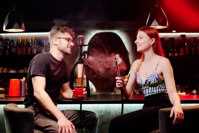 A man and woman sitting at a bar in a dimly lit lounge, enjoying shisha together. The man holds a red drink while both smile and relax in a stylish, modern setting.